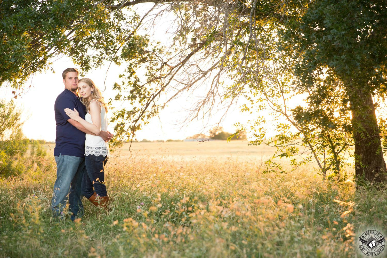 Wavy blond haired girl wearing a tank top with a lace fringe and blue jeans and boots softly hugs a sandy haired guy with a Van Dyke beard wearing a casual collared shirt with blue jeans under a green tree standing in a golden field of tall grass in Round Rock in this soft engagement image in a suburb or Austin, TX.